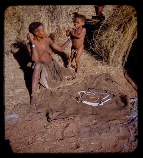 Di!ai (/Qui's wife) sitting and a child standing, with body ornaments displayed on a kaross on the ground in front of them