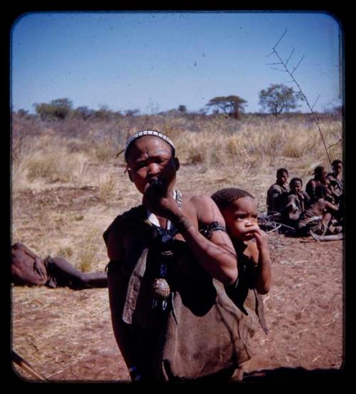 Di!ai (/Qui's wife) smoking, carrying a baby on her back, with a group of people sitting in the background