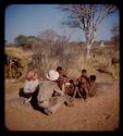 Group of boys sitting and making plasticene models, with a man cutting up meat behind them, Elizabeth Marshall Thomas and John Marshall watching