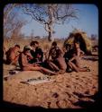 Group of boys sitting next to plasticene models of animals they made, with /Qui and ≠Toma cutting up meat in the background