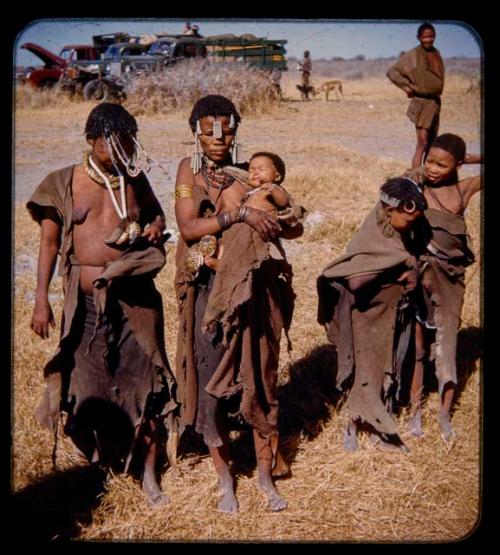 Four women standing, one holding a baby, with men, dogs and expedition trucks in the background