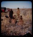 Group of people sitting, including /Qui, //Kushay, /Gao and !Ungka, with /Gao and Katambaye (an expedition member) standing next to them, expedition trucks in the background