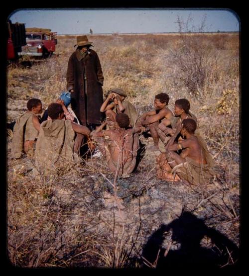 Group of people sitting, including /Qui, //Kushay, /Gao, !Ungka and /Gao, with Katambaye (an expedition member) standing next to them, expedition trucks in the background