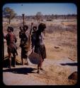 Head wife standing, holding a basket, with a group of women behind her beating mahangu, also known as pearl millet