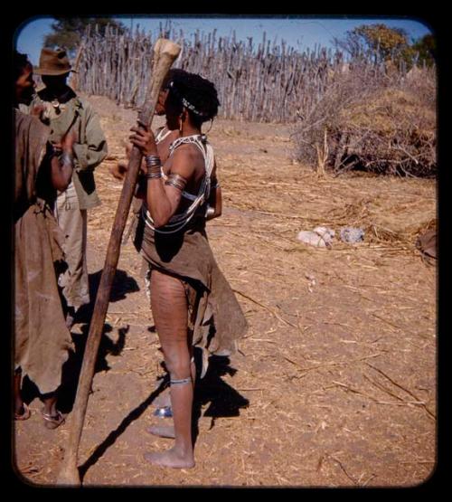 Woman with scarification on her leg, holding a pole for beating mahangu (pearl millet), standing with a group of people