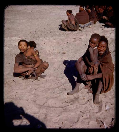 Two women sitting, one holding a child, the other with a child tied in her kaross