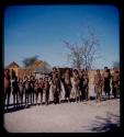 Group of people standing, with huts in the background