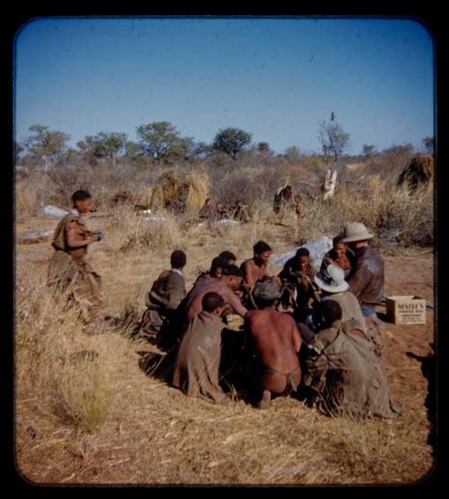 Group of people sitting, receiving mealies from an expedition member, with a woman walking toward them, people sitting in the background