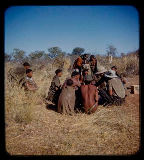 Group of people sitting, receiving mealies from an expedition member, with a woman walking toward them, people sitting in the background