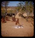 Group of women and children sitting near a cooking fire in Meri Catinga's kraal, with cattle, Kanakele's hut and another hut in the background