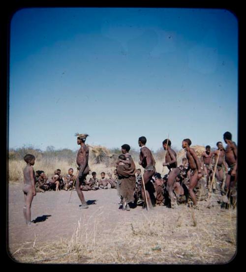 Dance: Group of men performing a curing dance around a circle of women, with a woman dancing holding a baby in her kaross