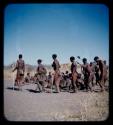 Dance: Group of men performing a curing dance around a circle of women, with a woman dancing holding a baby in her kaross