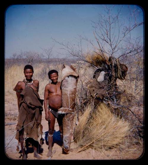 Possessions: Man holding rolled animal hide and a woman, standing by a tree hung with objects