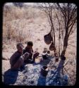 Skerm: Man and woman sitting by ashes and trees with possessions hanging from them
