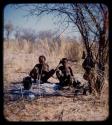 Skerm: Man and a woman sitting by ashes and trees with possessions hanging from them