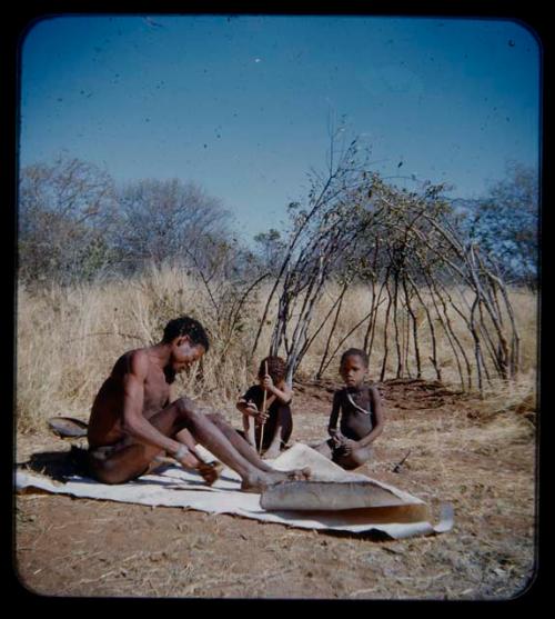 Skerm: Man sitting and working on a white animal hide, with two children sitting nearby