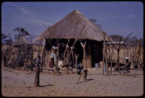 People outside a house, some sewing, seen through a fence