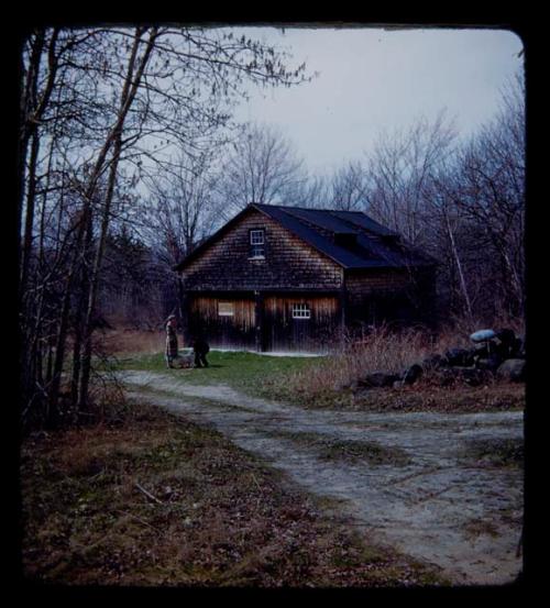 Lorna Marshall standing in front of a house, with another woman who is bending down to pet a dog, distant view