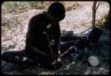 Man wearing a shirt, sitting under a tree, grinding something in a mortar