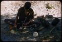 Man wearing a shirt, sitting under a tree, grinding something in a mortar