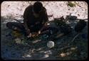 Man wearing a shirt, sitting under a tree, grinding something in a mortar