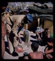 Expedition: Expedition member bending over to pour water from a kettle into an ostrich eggshell held by a seated man, with other people watching