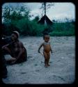 Expedition: Boy standing, with a woman with leg scarifications sitting nearby and a tent in the background