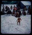 Expedition: Boy standing, with a group of people, including Lorna Marshall, sitting by a tent in the background