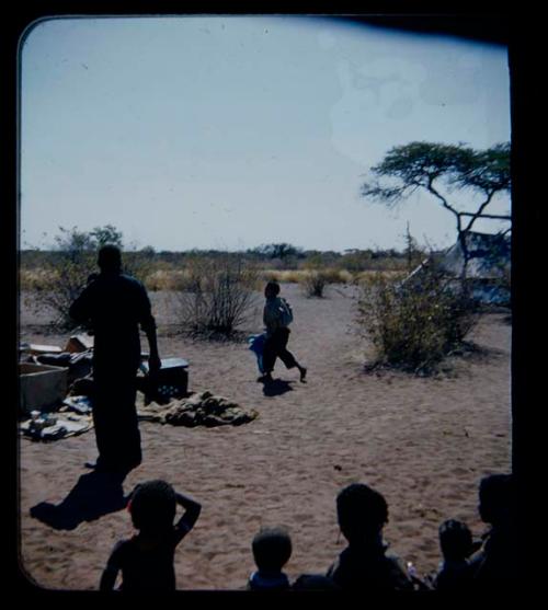 Expedition: Women and children sitting, with one of the expedition members and a boy wearing a large shirt and pants walking in the background