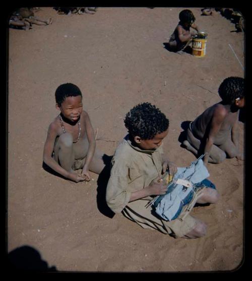 Expedition: Four children sitting; one boy in the foreground is holding bundle of clothing and another boy in the background is playing with a tin can