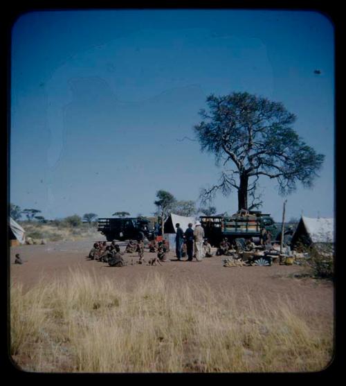 Expedition: Lorna Marshall,  Laurence Marshall, and Kernel Ledimo handing out gifts to people, with a tent and a truck with other expedition members in the background