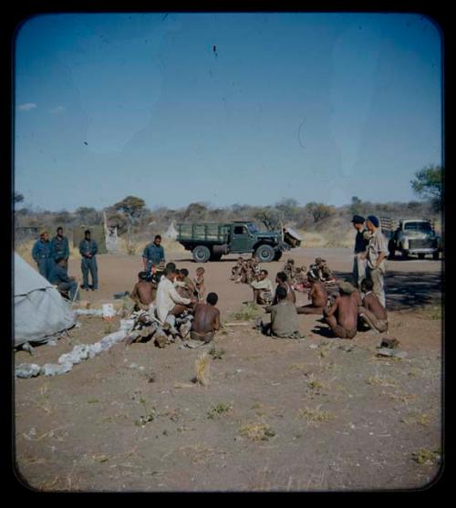 Expedition: Group of people sitting and expedition members standing near them, with trucks in the background