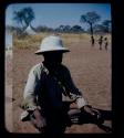 Expedition: ≠Toma wearing Western clothes (a shirt and a helmet) and sitting, with three children standing in the background