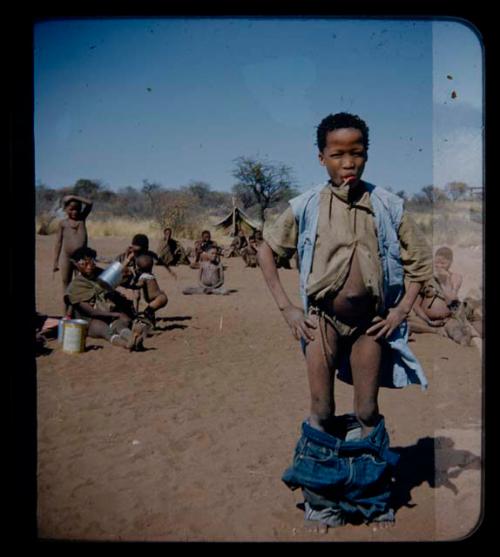 Expedition: Boy with a lollipop in his mouth, wearing two shirts and jeans which have fallen, with a group of people in the background