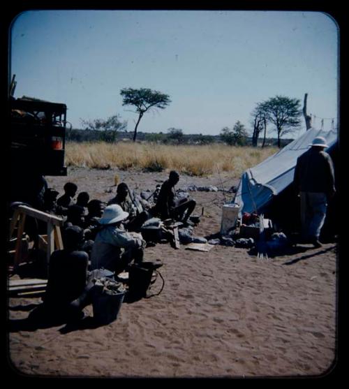 Expedition: Group of people sitting by a truck with gifts and Laurence Marshall walking into a tent nearby