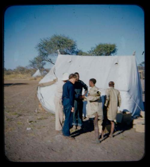 Expedition: Laurence Marshall shaking hands with a man wearing a shirt, with Lorna Marshall, Kernel Ledimo, and a man standing, near a tent