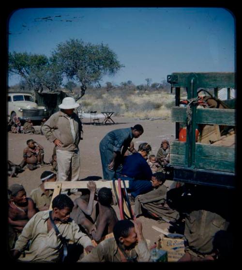 Expedition: Lorna Marshall and Kernel Ledimo probably getting gifts ready near a truck, with Laurence Marshall standing behind them and a group of people sitting in the background