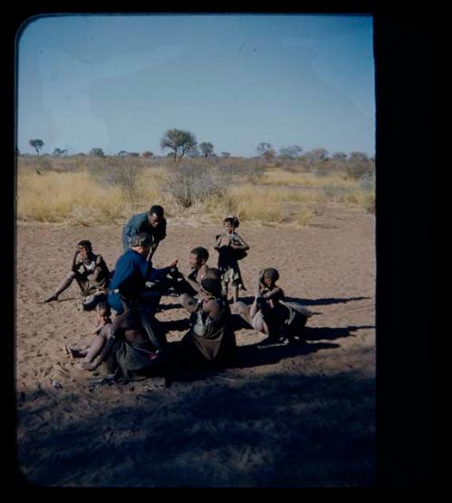 Expedition: Lorna Marshall giving something to an elderly man, with Kernel Ledimo present and a group of people sitting nearby