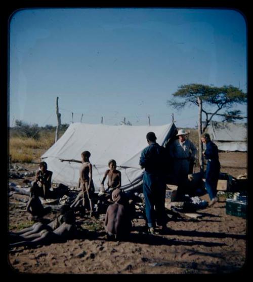 Expedition: Laurence Marshall, Lorna Marshall, and Kernel Ledimo standing in front of a tent, with a group of people sitting with gifts nearby