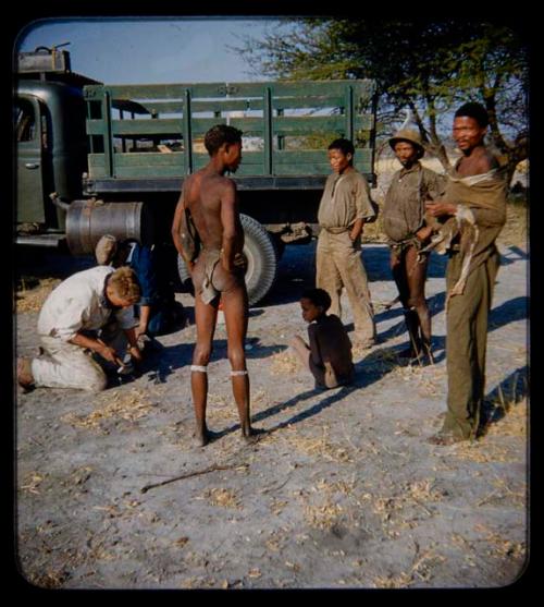 Expedition: Group of men, including ≠Toma, standing and a child sitting by a truck; John Marshall and another expedition member repairing something nearby