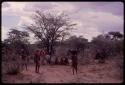 Three women walking past a group of people sitting, being interviewed by Lorna Marshall, with another expedition member interpreting