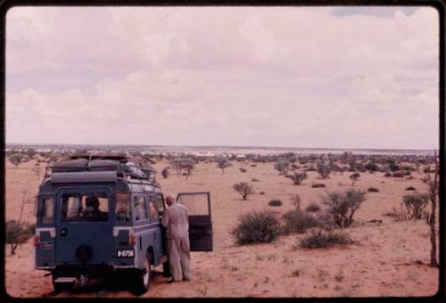 Laurence Marshall standing next to the expedition Land Rover, view from behind, with a pan in the distance