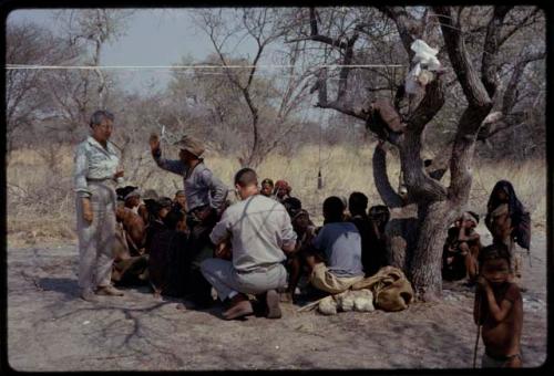 Group of people sitting with Ngani and Nicholas England in the expedition camp under a clothesline, with Lorna Marshall standing next to them