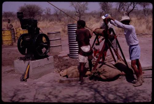 Group of people building a water pump; ǂToma and /Gao pulling on a rope and Kernel Ledimo kneeling by the cement base