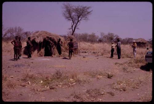 Group standing before ǂToma's hut, includes Lorna Marshall, Wilhelm Camm, and Ngani