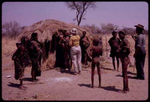 Group standing before ǂToma's hut, includes Lorna Marshall, Wilhelm Camm, and Ngani