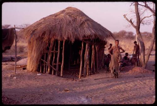 /Naoka, /Ti!kay's first wife, standing in front of /Ti!kay's hut