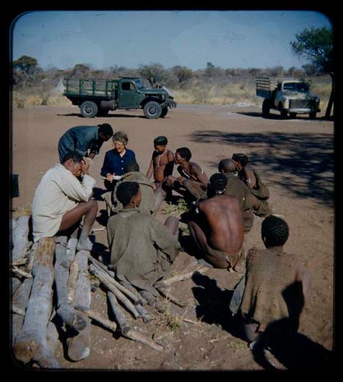 Group of people being interviewed by Lorna Marshall and another expedition member, with trucks in the background