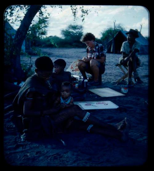 Expedition: Women and children with drawings and painting tools, with Elizabeth Marshall Thomas marking a completed painting and Ngani sitting in a chair next to her in the background