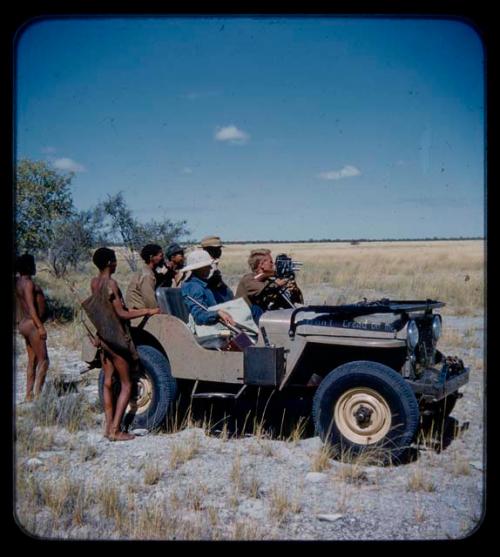 Expedition: John Marshall holding a camera, with Lorna Marshall, Ngani, and other expedition members riding in Jeep with him; two men with hunting equipment are standing nearby